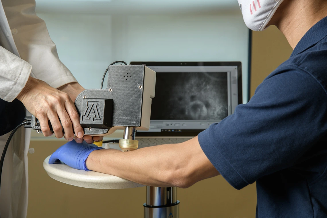 Clara Curiel Lewandrowski, MD, uses a portable skin cancer diagnostic microscope on Dongkyun Kang, PhD, to test the output image on a laptop screen. The two are developing the device to enable faster skin cancer diagnosis in doctor’s offices.