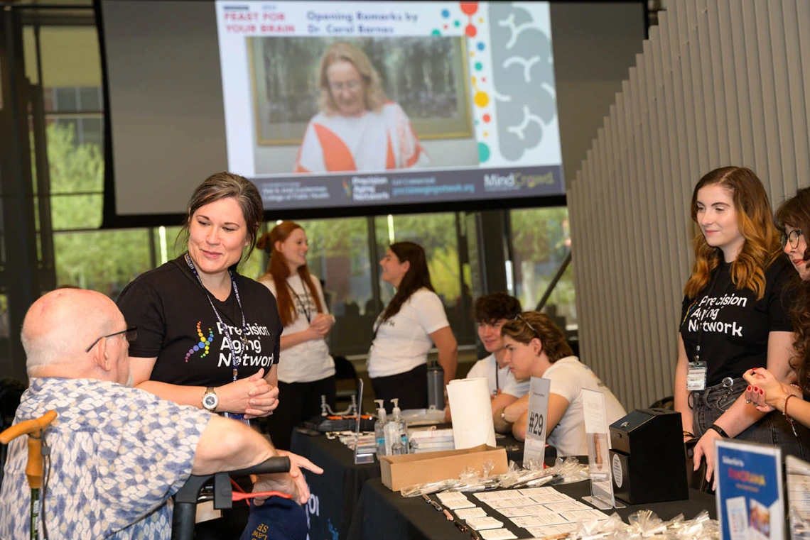 Two women wearing t-shirts that read "Precision Aging Network" talk with an older man in a wheelchair inside a large open room with other people in the background. 