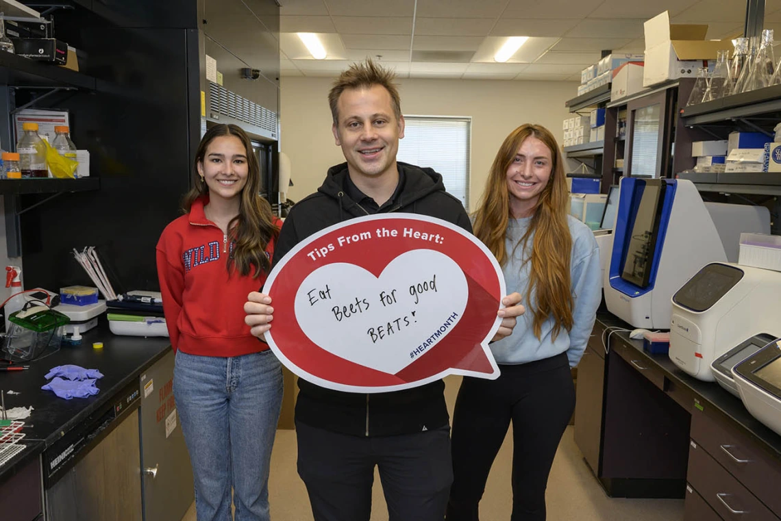 White man in dark hoody stands in lab holding a heart poster with two female students standing slightly behand him.