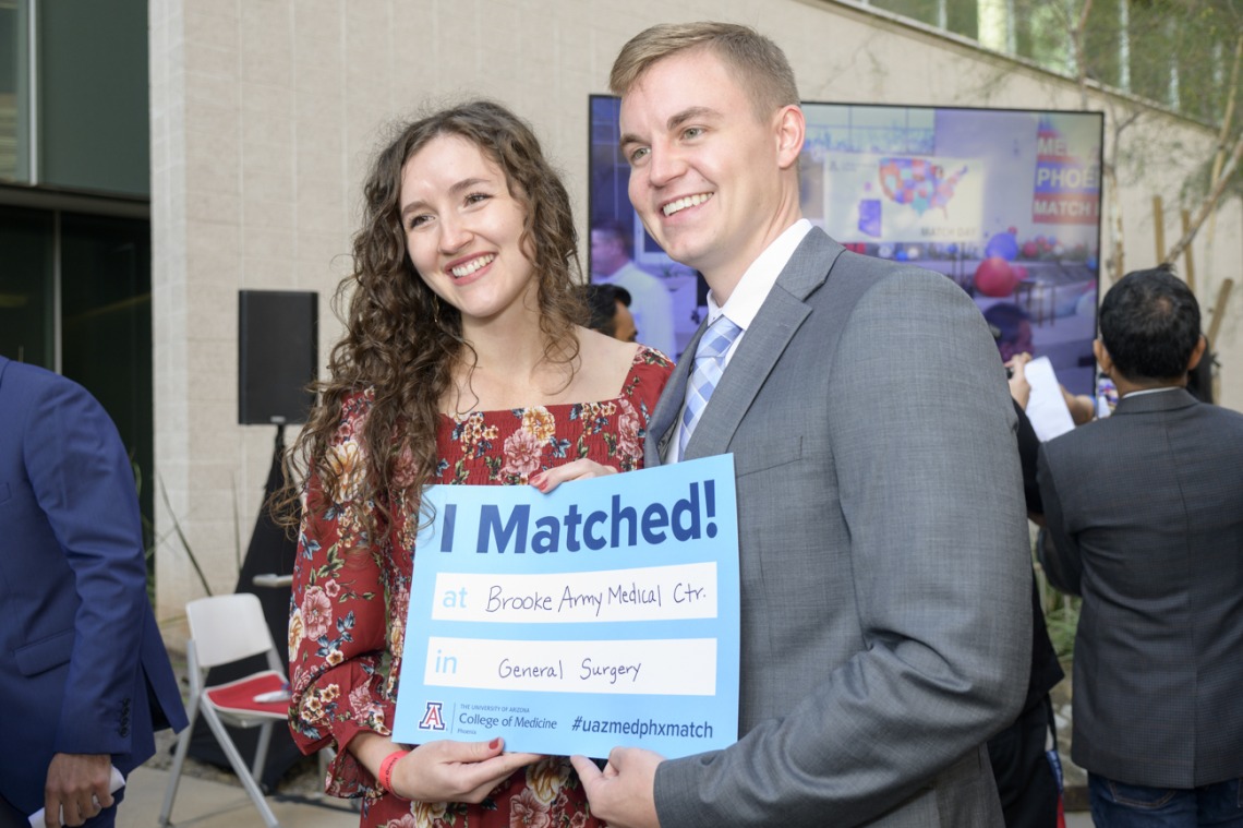 A young adult, light skinned man and woman stand together smiling and holding a Match Day poster.