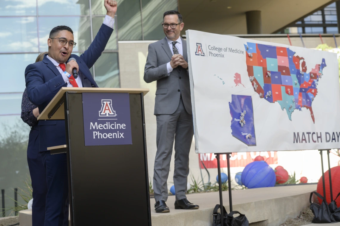 A young adult male wearing a suite and tie raises a hand in celebration while standing at a podium. A map of the US is in the background. 