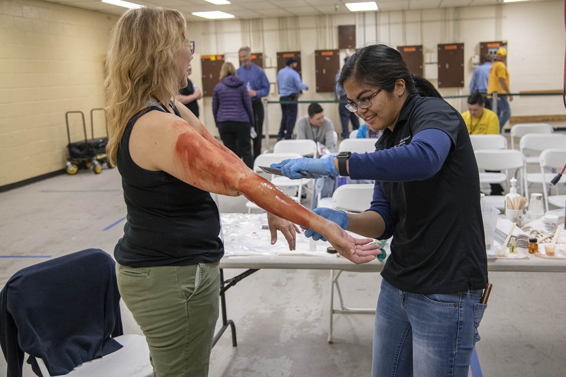 With outstretched arms, volunteer Pamela Leonard prepares to roleplay an airplane passenger injured by a midair explosion. Merryl Lopido, simulation operations specialist for ASTEC, uses special-effects makeup to create fake burns.