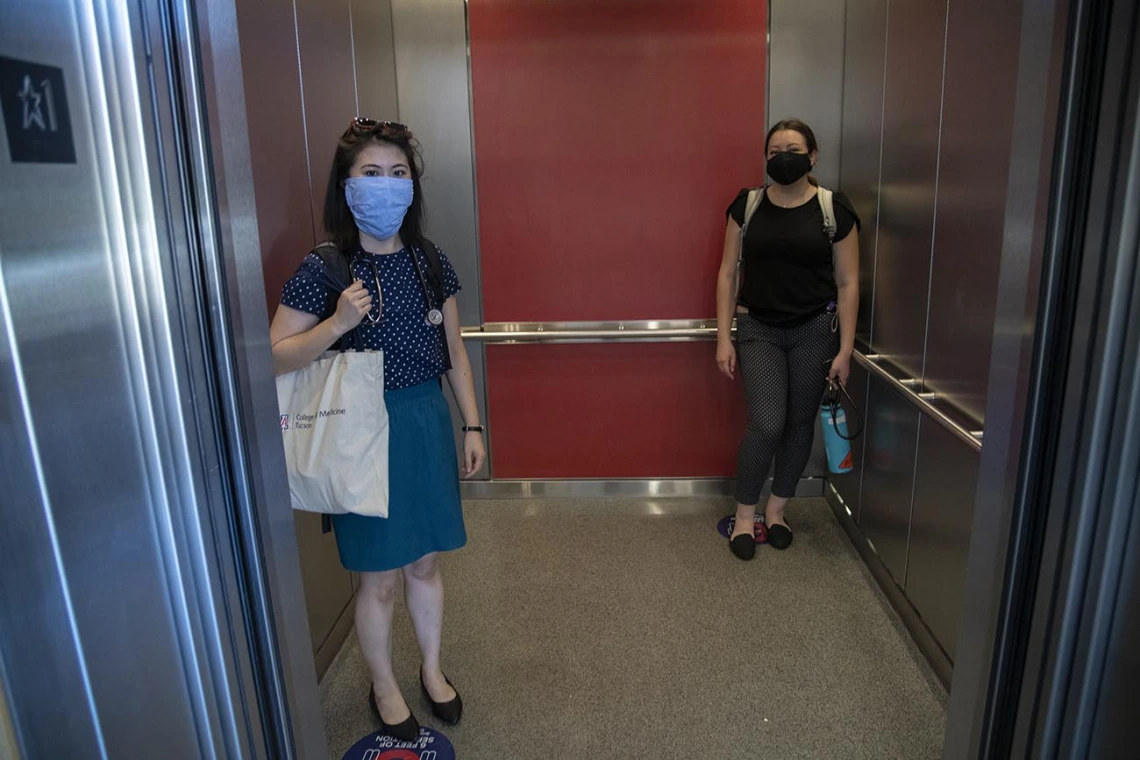 First-year College of Medicine – Tucson students Aileen Lee (left), and Brianne Davis (right) stand on signs placed on elevator floors marking six feet.