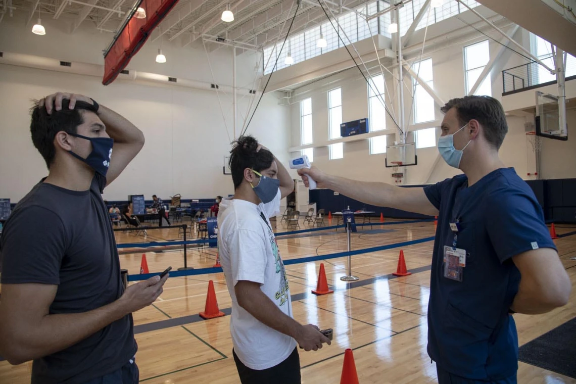 University of Arizona student Heath Zuniga (left) watches as Wyatt Snell, a clinical research coordinator, takes student Mason Young’s temperature prior to the self-administered COVID-19 testing procedure.