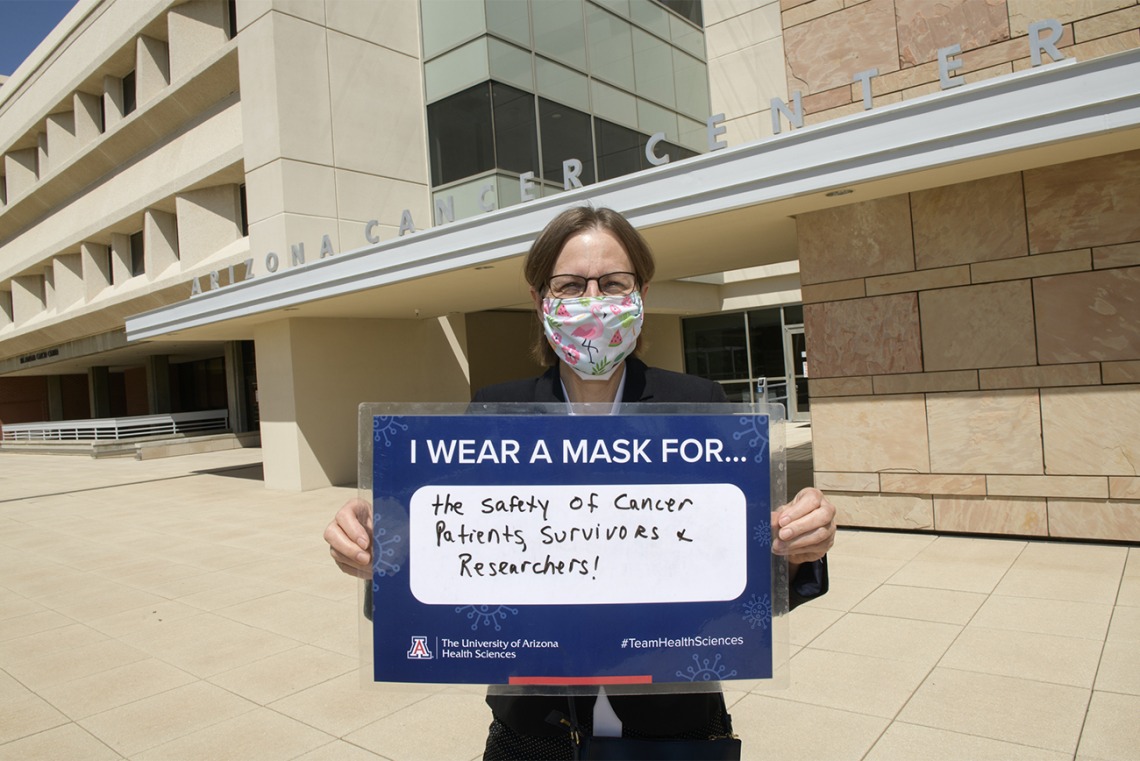 UArizona Cancer Center director Joann Sweasy, PhD, holds her sign in front of the Cancer Center’s Levy and Salmon buildings  in Tucson. She wears a mask for “the safety of cancer patients, survivors and researchers.”