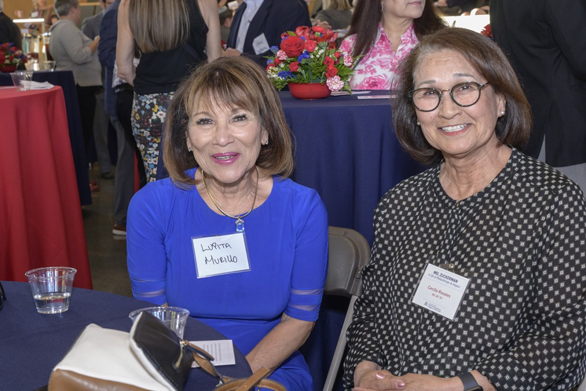 Two older women with shoulder-length brown hair sit at a table smiling.