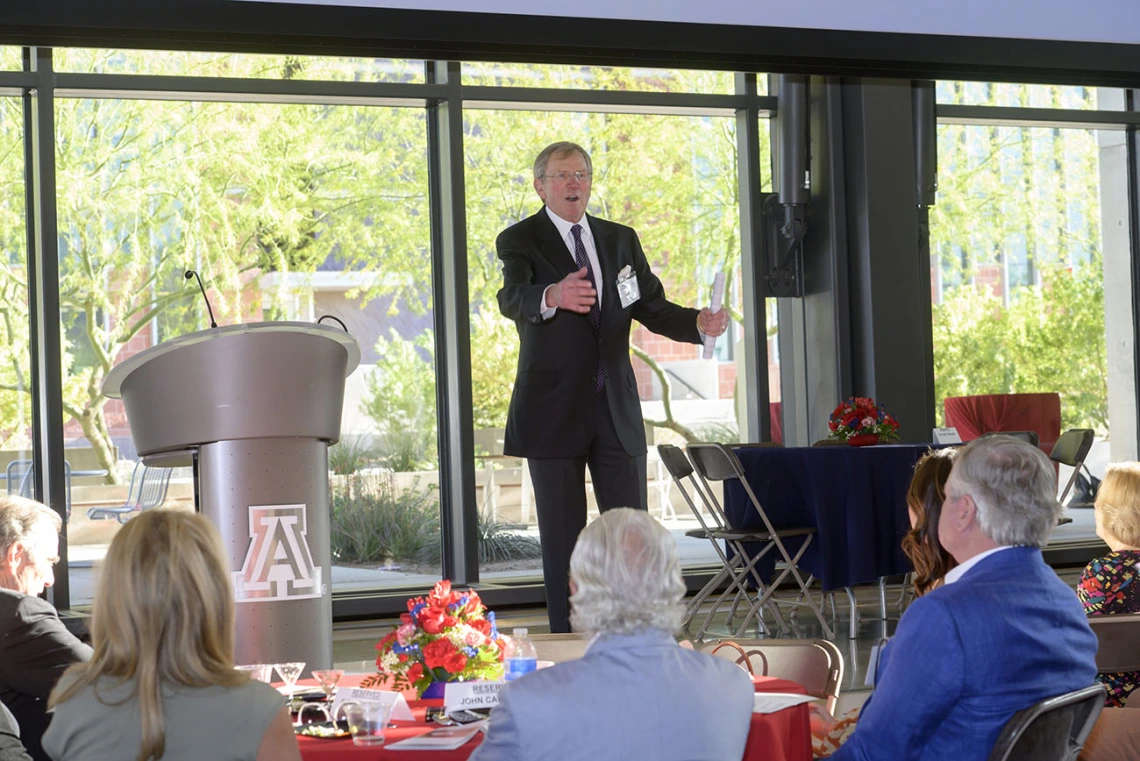 An older light-skinned man in a suite stands on a stage talking to a crowd. 