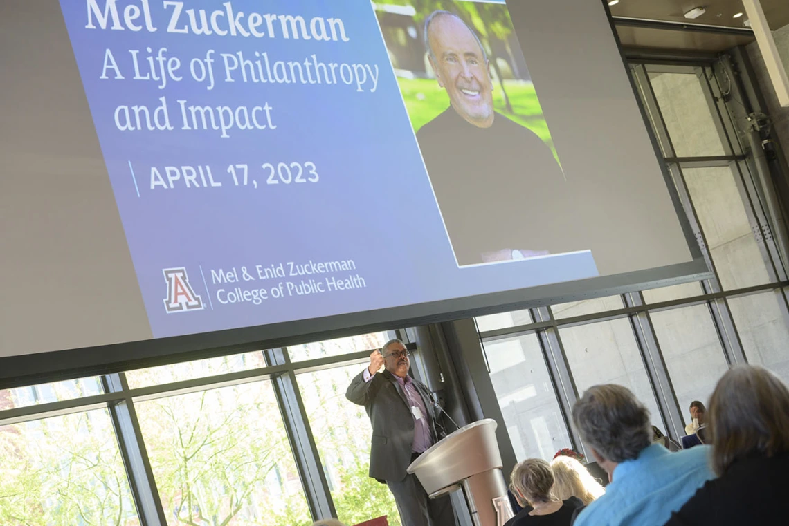 A tall dark-skinned man in a suite stands at a podium jesturing as he speaks under a large video display of Mel Zuckerman.