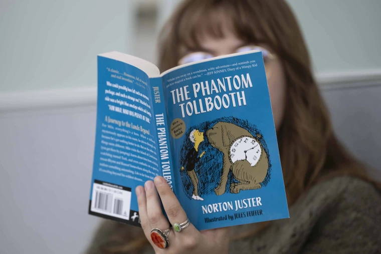 Close-up photo of the book “The Phantom Tollbooth” being help up and read by a person who is blurred into the background