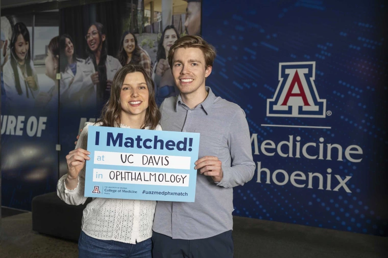 Kate Chartrand stands next to husband Nick Chartrand in front of the U of A College of Medicine – Phoenix sign. Both are holding an “I Matched!” sign that reads UC Davis and ophthalmology beneath it.