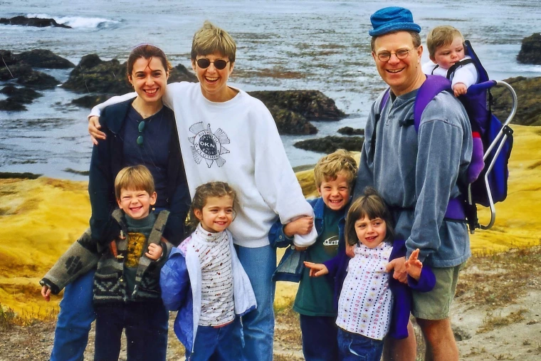 A young woman stands next to another woman who has her arm around her while a man wearing a small blue hat has a baby in a backpack on his back. Four small children stand in front of them, smiling.  