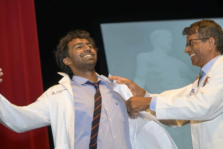 A new medical student laughs as a smiling professor helps him put on his white coat. 