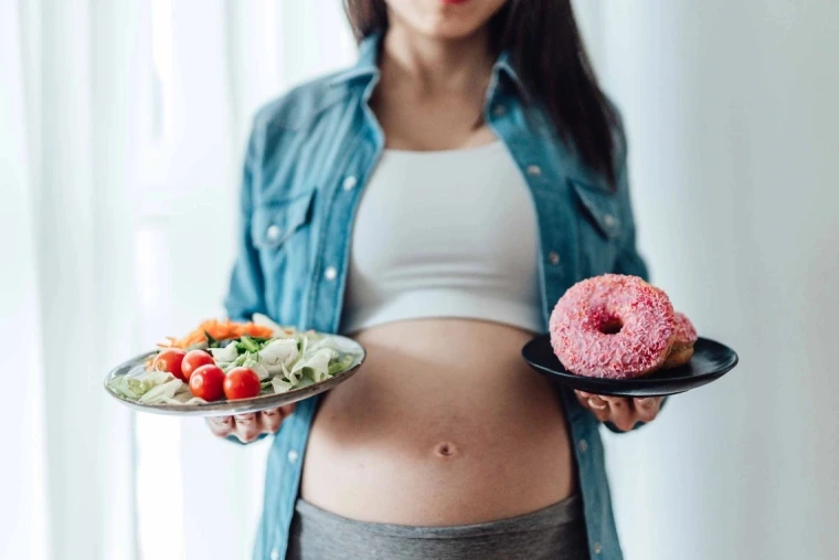 A pregnant woman, whose belly is showing, holds two plates of food in her outstretched hands. On one plate is a salad. On the other plate are a couple of large doughnuts.