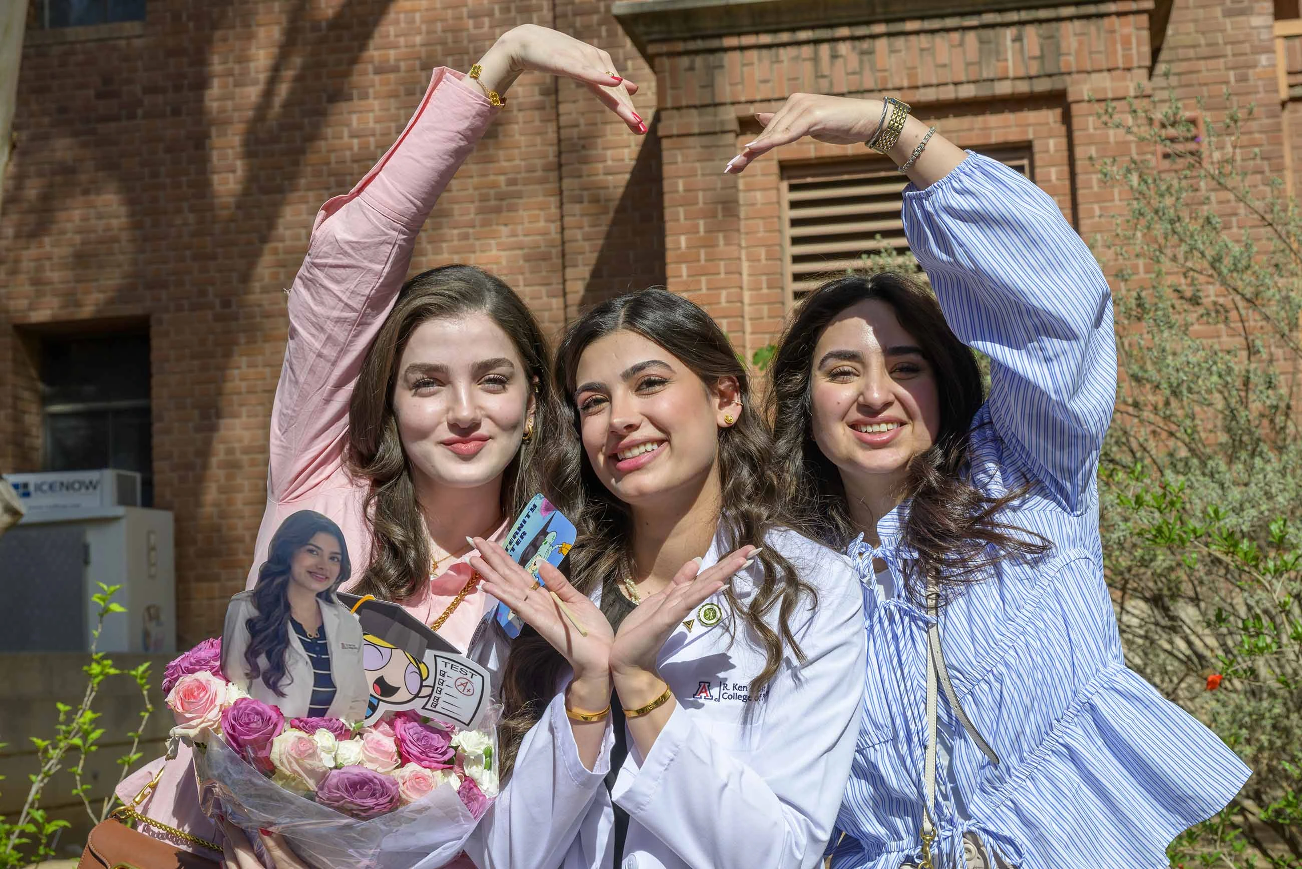 Three smiling young women create a heart shape with their arms. One of them is wearing a medical white coat. 