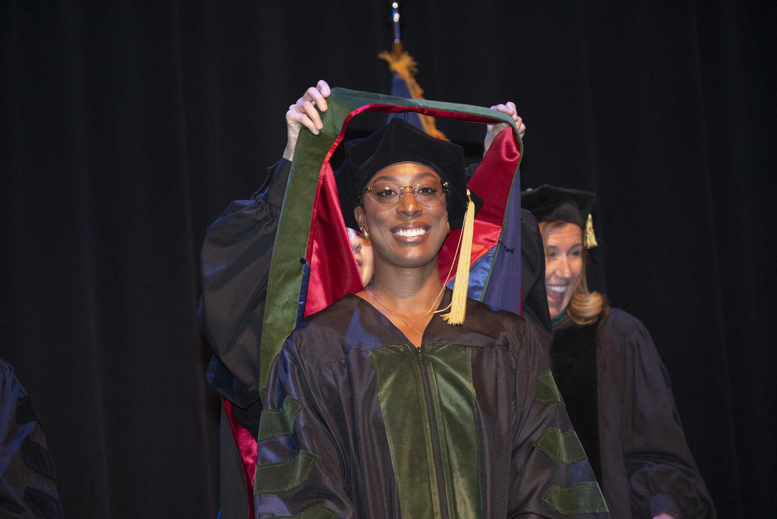 A smiling woman wearing graduation regalia looks ahead as someone behind her places a ceremonial hood over her shoulders. 