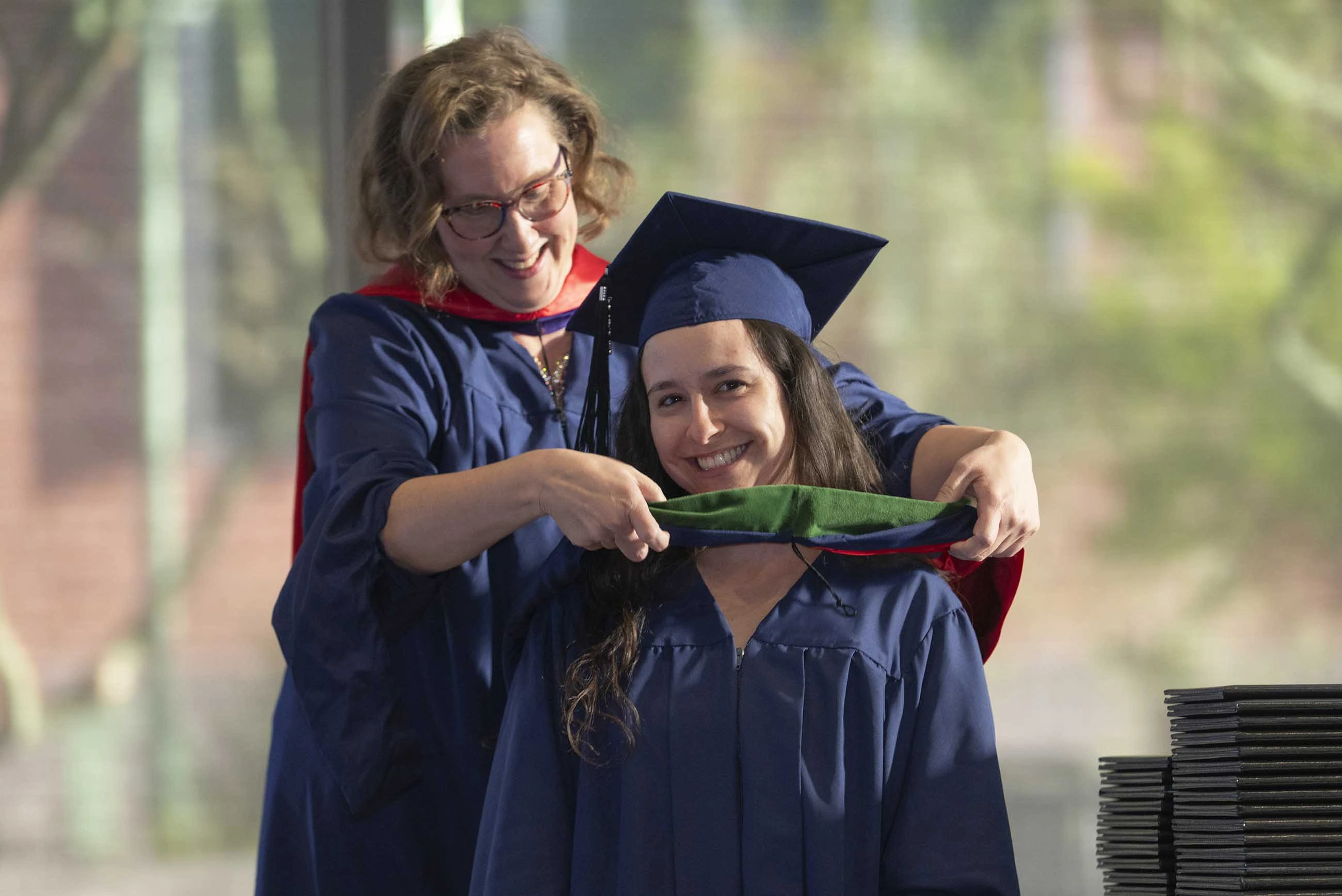 A professor places a ceremonial hood over the shoulders of a young woman. Both are smiling and wearing graduation regalia. 