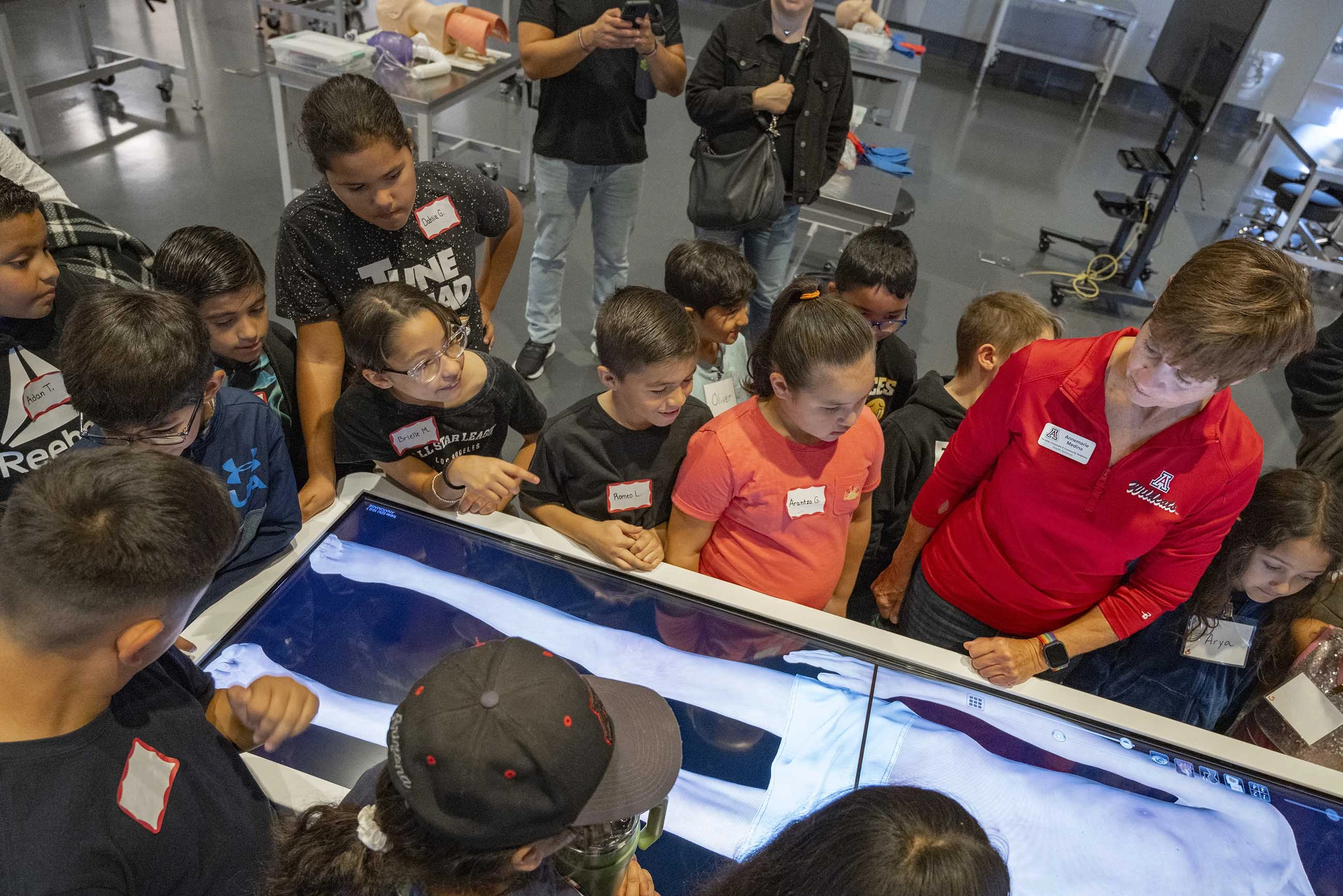 About a dozen grade-school students stand around a table with a digital image of a body while a tour guide demonstrates how it works. 