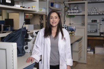 woman with long brown hair wearing a white lab coat, standing in research lab