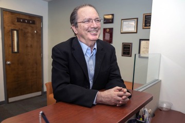 A smiling man wearing glasses stands at a desk with his hands folded together.