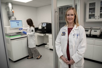 Dr. Typpo, a woman with medium length blonde hair, wearing a red blouse under a white lab coat with a Steele Children's Research Center patch. She is standing in a lab in front of the new Nova Seq 600 genome sequencer.