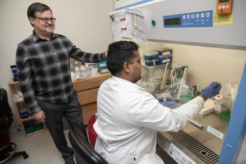 Dr. Rafikov, with dark hair and glasses wearing a black and white checkered shirt, stands watching Joel James (a lab technician) sitting at a lab station, preparing a sample for sequencing. The technician is wearing a white lab coat.