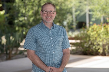 A portrait of Dr. Edwards, a man with brown hair, glasses and a green button-up short sleeved shirt, standing outside.