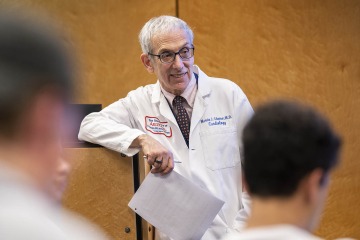 Older white male wearing glasses and a white coat stands at the front of a class giving a lecture to students.