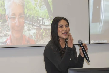 A woman stands in front of a projector screen holding a microphone and gesticulating while speaking.