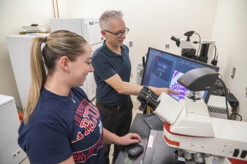 Dr. Frank von Hippel, a middle-aged white man stands at a lab table pointing to a moniter while talking with a young white woman in front of a microscope. 