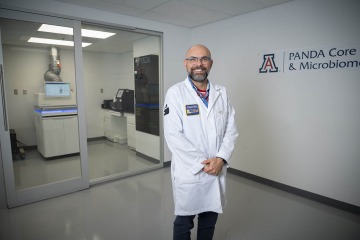 Dr. Laubitz, a man with facial hair and shaved head wearing a white lab coat, stands in front of the PANDA Core and Microbiome lab.