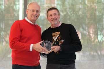 Older man in red sweater standing with man in dark sweater with Christmas bear on it holding glass pharmacy award.