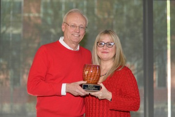 Tall older man in red sweater standing with shorter woman with blond hair in red sweater holding glass award.