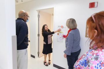 A woman with dark hair points to signage outside an exam room during a tour with several people looking on. 