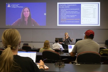 Jenene Spencer, PharmD, instructs a classroom of pharmacy students while Sandi Thoi, PharmD, appears on a projector screen via Zoom.
