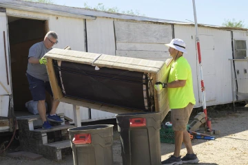 Two men carry a couch out of a run-down trailer.