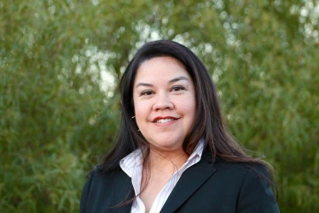 Woman with long dark hair stands smiling outside. She is wearing a black blazer and white dress shirt.