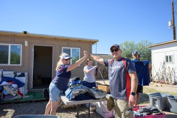 A woman with long blonde hair wearing a baseball cap gives a high-five to a man in a baseball hat as their arms stretch out above a table covered in donated cloting. 