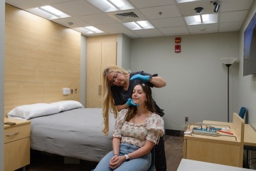 In a bedroom setting a woman stands behind another seated woman and attaches electrodes to her head in preparation for a sleep study. 