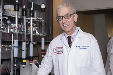 Older man wearing white coat stands in a lab setting