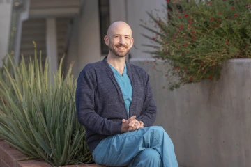 Man wearing scrubs sits outside.