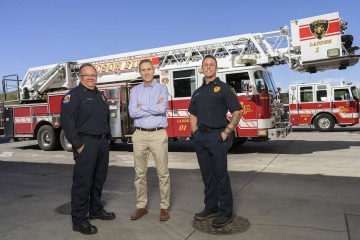 Two firefighters and another man stand in front of a fire truck.