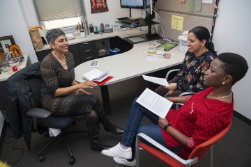 South Asian professor sits at her desk talking with two of her mentees.
