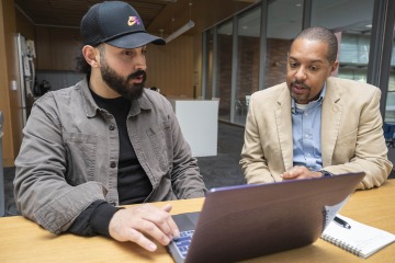 Photo of male student in a baseball cap looking at a laptop computer and sitting next to Michael D.L. Johnson. 