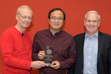 Three men stand next to each other holding a glass award between them, all smiling.
