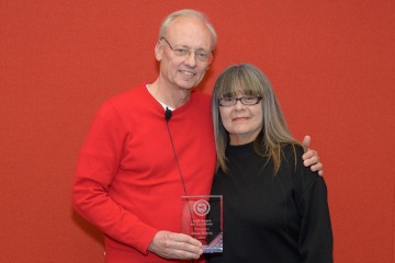 A woman and man stand next to each other holding a glass award between them, both smiling.