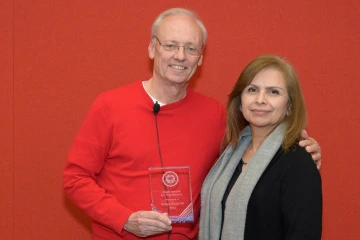 A woman and man stand next to each other holding a glass award between them, both smiling.