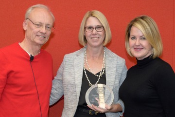 A man and two women stand next to each other holding a glass award between them, all smiling.