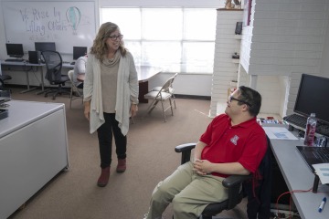 Lupita Loftus talks with Gabe Martinez, who’s sitting in a chair at his desk.