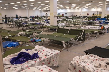 A large room is filled with green cots covered in American Red Cross blankets.