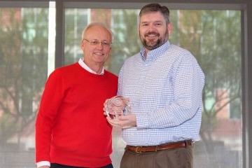 Slender older man in red sweater standing with a taller, younger bearded man in blue shirt holding award.
